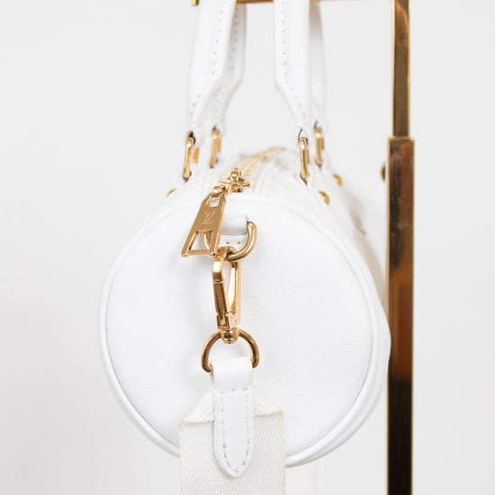 A white round shoulder bag with gold-tone hardware and a white canvas strap, hanging against a white background with a gold metal display stand visible on the right.