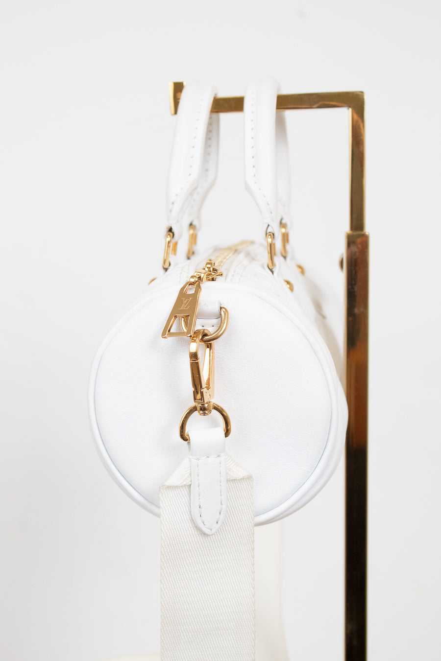 A white round shoulder bag with gold-tone hardware and a white canvas strap, hanging against a white background with a gold metal display stand visible on the right.
