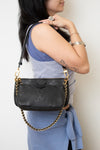 A woman wearing a light blue top carrying a black Louis Vuitton shoulder bag with an embossed monogram pattern and a gold-tone chain strap.