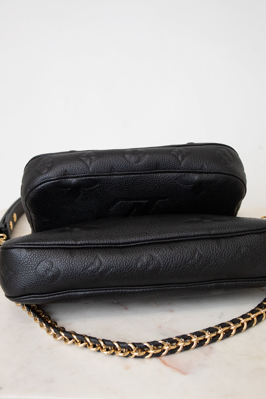 Underside of a black leather shoulder bag with gold-tone hardware and a chain strap, against a light gray background.