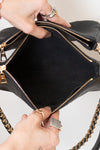 Hands holding open a purse showing a black interior of a black leather shoulder bag with gold-tone hardware and a chain strap, against a light gray background.