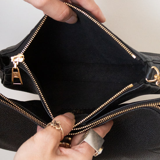 Close-up of a black handbag being opened by two hands, showing the interior lining and gold-tone zipper hardware against a light neutral background.