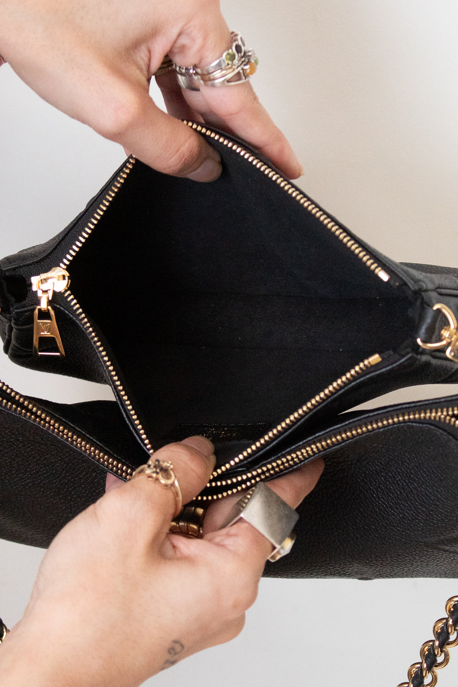 Close-up of a black handbag being opened by two hands, showing the interior lining and gold-tone zipper hardware against a light neutral background.