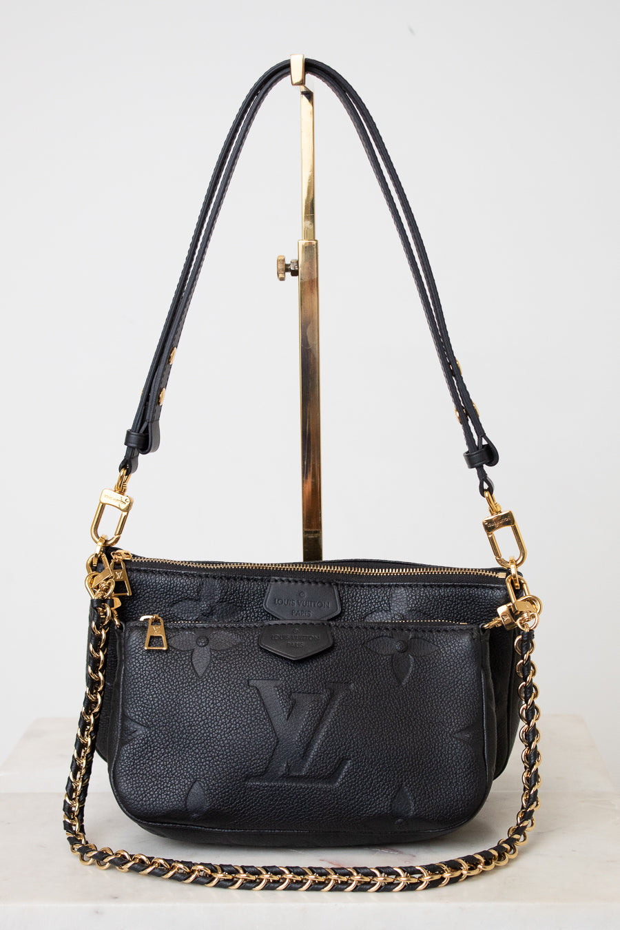 A black Louis Vuitton monogram shoulder bag with gold-tone hardware and a gold chain strap, displayed on a white background with a gold stand.