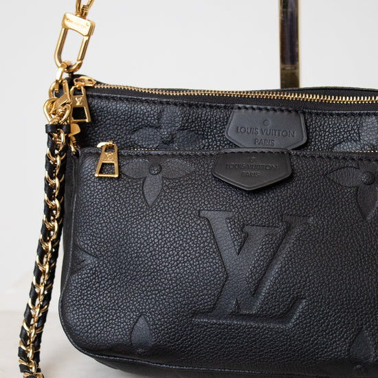 A close-up of a black Louis Vuitton shoulder bag with a pebbled leather texture, embossed monogram pattern, gold-tone hardware, and a black-and-gold chain strap on a light marble surface.