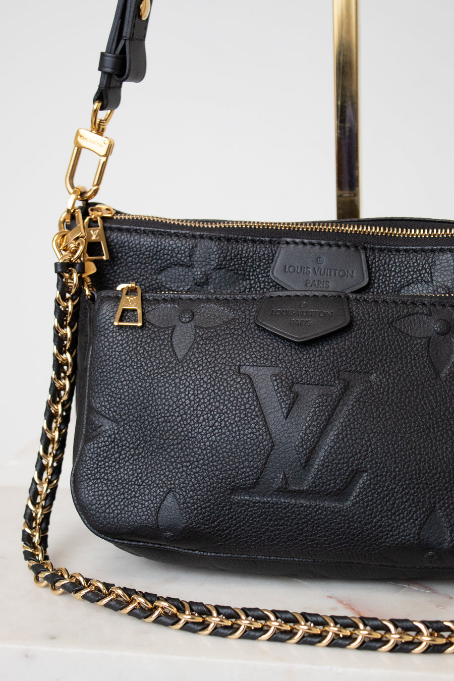 A close-up of a black Louis Vuitton shoulder bag with a pebbled leather texture, embossed monogram pattern, gold-tone hardware, and a black-and-gold chain strap on a light marble surface.
