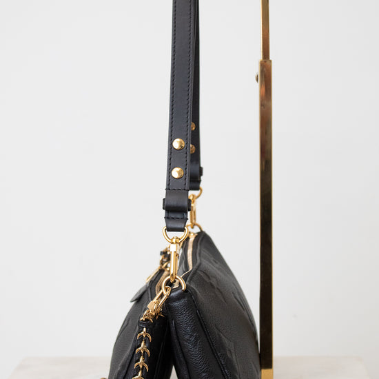 A black leather shoulder bag with gold-tone hardware and a gold chain strap, displayed on a minimalist gold metal stand against a clean white background.