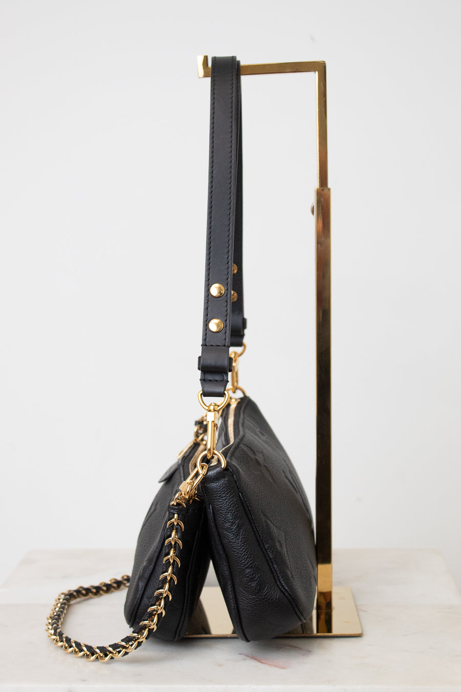 A black leather shoulder bag with gold-tone hardware and a gold chain strap, displayed on a minimalist gold metal stand against a clean white background.