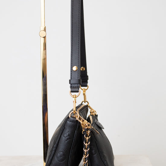 A black leather shoulder bag with gold-tone hardware and a chain strap, displayed on a  gold metal stand against a light gray background.