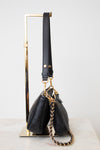 A black leather shoulder bag with gold-tone hardware and a chain strap, displayed on a  gold metal stand against a light gray background.
