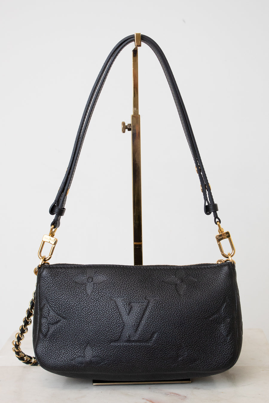 A black leather shoulder bag with gold-tone hardware and a chain strap, displayed on a  gold metal stand against a light gray background.