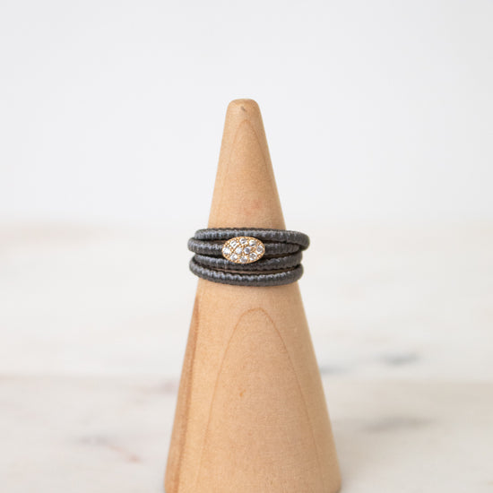 A stack of three dark gray braided rings with a central oval cluster of small clear stones, displayed on a light wood cone stand against a soft white background.