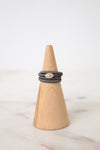 A stack of three dark gray braided rings with a central oval cluster of small clear stones, displayed on a light wood cone stand against a soft white background.