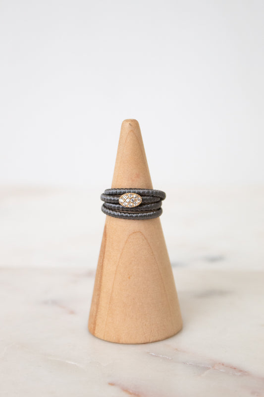 A stack of three dark gray braided rings with a central oval cluster of small clear stones, displayed on a light wood cone stand against a soft white background.