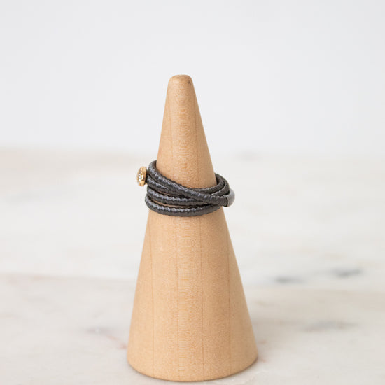A dark gray multi-strand wrap ring with a small gold bead accent, displayed on a light wood cone jewelry stand over a white marble surface.