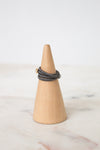 A dark gray multi-strand wrap ring with a small gold bead accent, displayed on a light wood cone jewelry stand over a white marble surface.