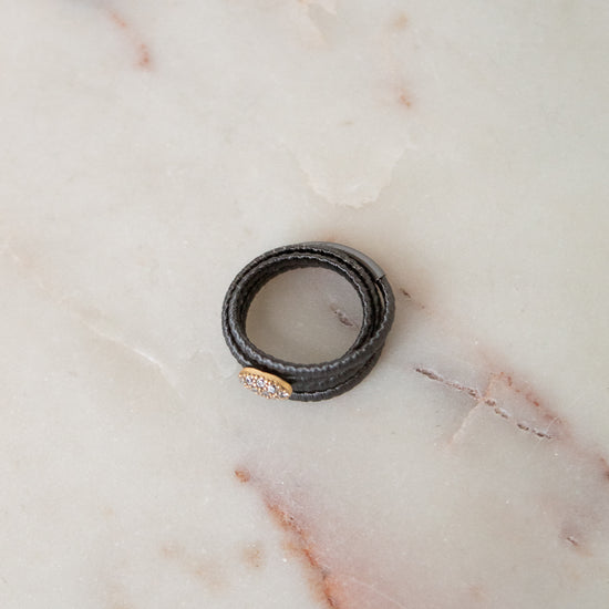 A black textured leather wrap ring with a small gold oval charm, resting on a light beige marble surface.