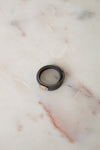 A black textured leather wrap ring with a small gold oval charm, resting on a light beige marble surface.