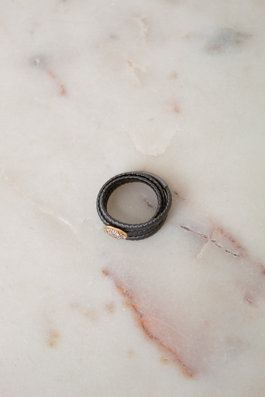 A black textured leather wrap ring with a small gold oval charm, resting on a light beige marble surface.