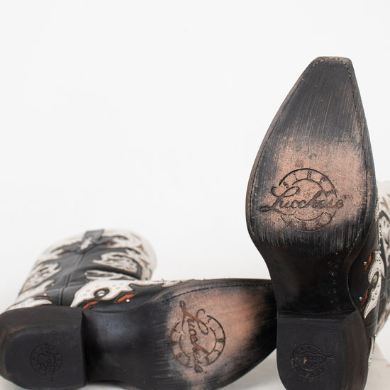A pair of cowboy boots shown from the sole side, featuring a black leather heel and toe cap with a circular logo, and a white leather with black western-style patterning.