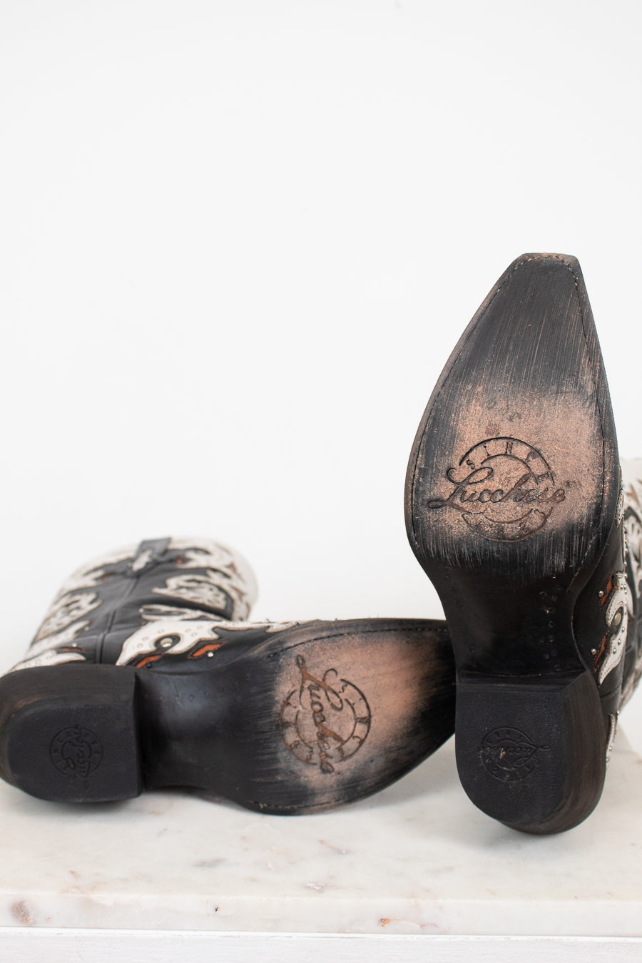 A pair of cowboy boots shown from the sole side, featuring a black leather heel and toe cap with a circular logo, and a white leather with black western-style patterning.