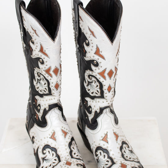 A pair of western cowboy boots with a black and white leather design, featuring intricate brown and black embroidery and silver-tone studs, displayed on a white marble surface against a white background.