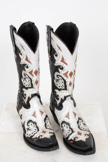 A pair of western cowboy boots with a black and white leather design, featuring intricate brown and black embroidery and silver-tone studs, displayed on a white marble surface against a white background.