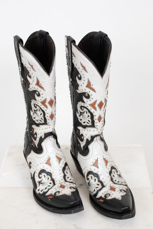 A pair of western cowboy boots with a black and white leather design, featuring intricate brown and black embroidery and silver-tone studs, displayed on a white marble surface against a white background.