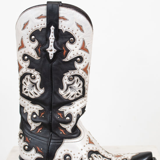 A single western cowboy boot with a black and white leather base, featuring intricate orange-brown accents, and silver studded detail, set against a plain white background.