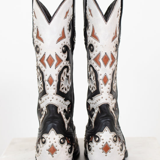 A pair of western cowboy boots shown from the back, featuring a black and white leather base with intricate brown and black leather detailing and silver studs.