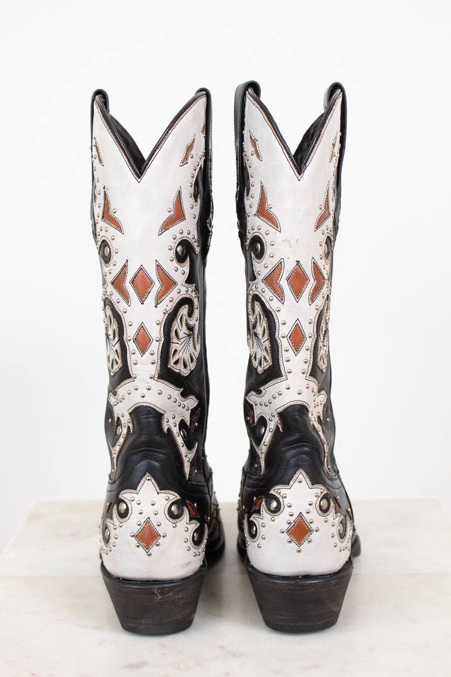 A pair of western cowboy boots shown from the back, featuring a black and white leather base with intricate brown and black leather detailing and silver studs.