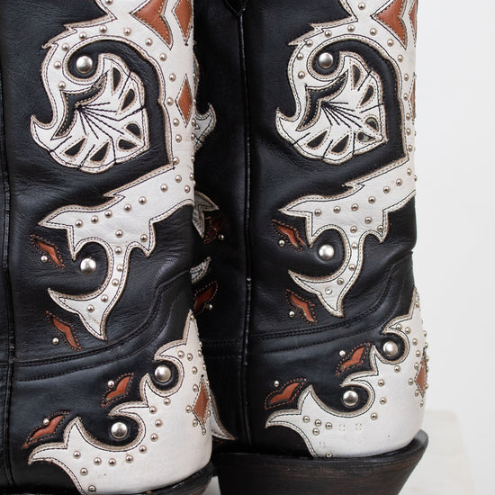 Close-up of the heel of a pair of western cowboy boots in black leather with intricate white and brown details, silver studs, and dark brown stacked heels.