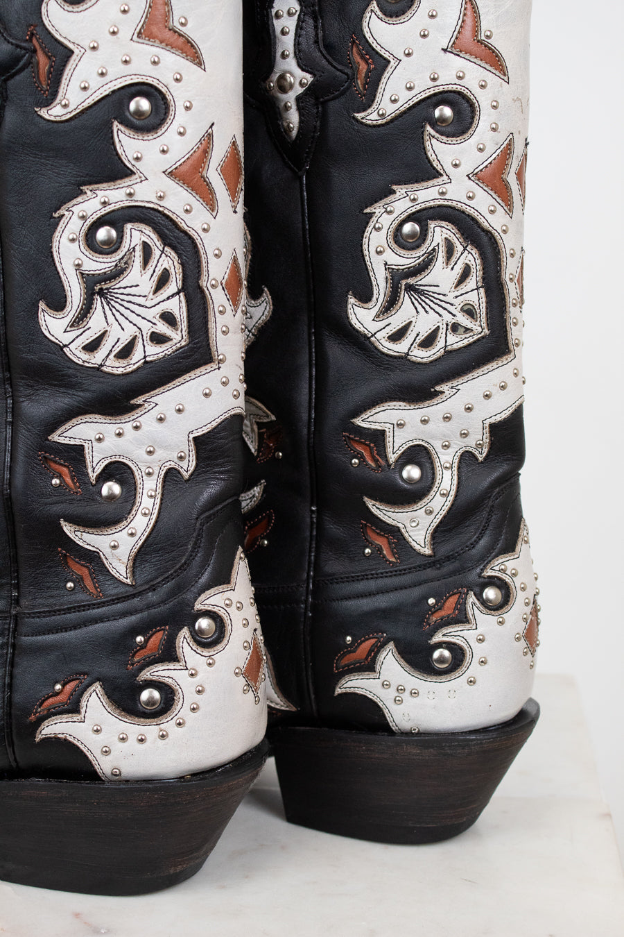 Close-up of the heel of a pair of western cowboy boots in black leather with intricate white and brown details, silver studs, and dark brown stacked heels.