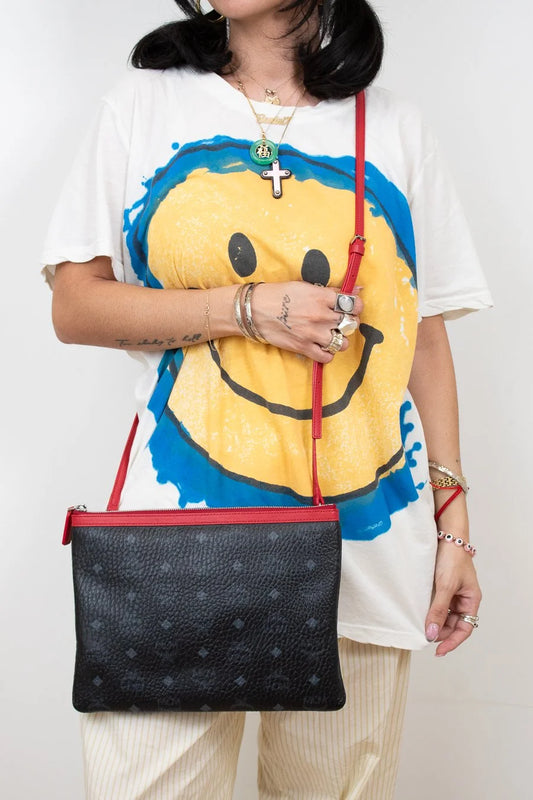 Person wearing a white t-shirt with a smiley face design, holding a black handbag with red straps.