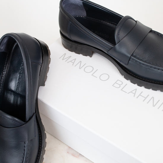 A pair of navy leather penny loafers with chunky black soles, displayed on a white marble surface next to a white shoebox printed with the brand name 'MANOLO BLAHNIK'.