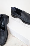 A pair of navy leather penny loafers with chunky black soles, displayed on a white marble surface next to a white shoebox printed with the brand name 'MANOLO BLAHNIK'.