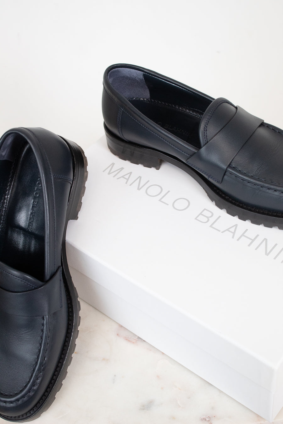 A pair of navy leather penny loafers with chunky black soles, displayed on a white marble surface next to a white shoebox printed with the brand name 'MANOLO BLAHNIK'.