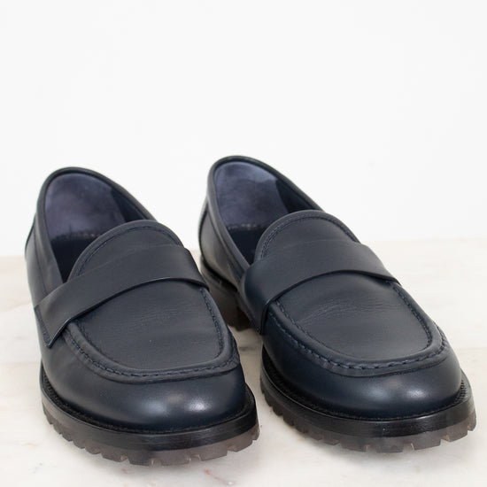 A pair of dark navy blue leather penny loafers with thick black lug soles, displayed on a light marble surface against a white background.