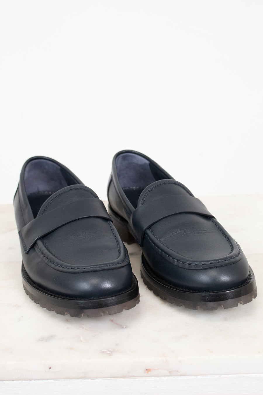 A pair of dark navy blue leather penny loafers with thick black lug soles, displayed on a light marble surface against a white background.
