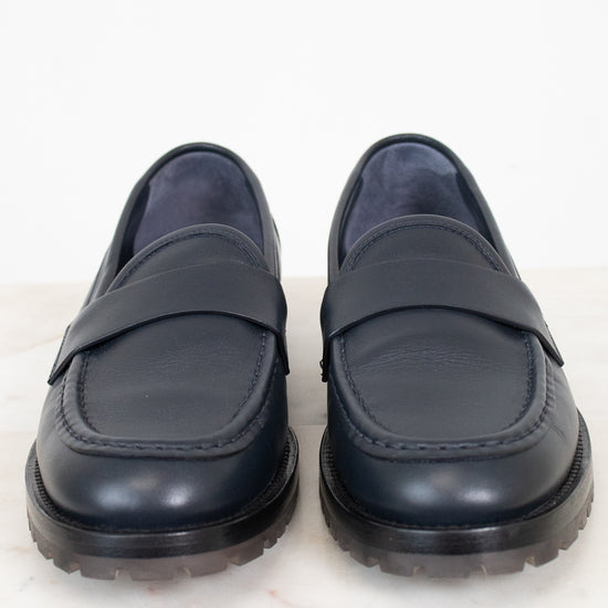 A pair of navy leather penny loafers with thick black soles, displayed side-by-side on a white marble surface against a white background.