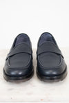 A pair of navy leather penny loafers with thick black soles, displayed side-by-side on a white marble surface against a white background.