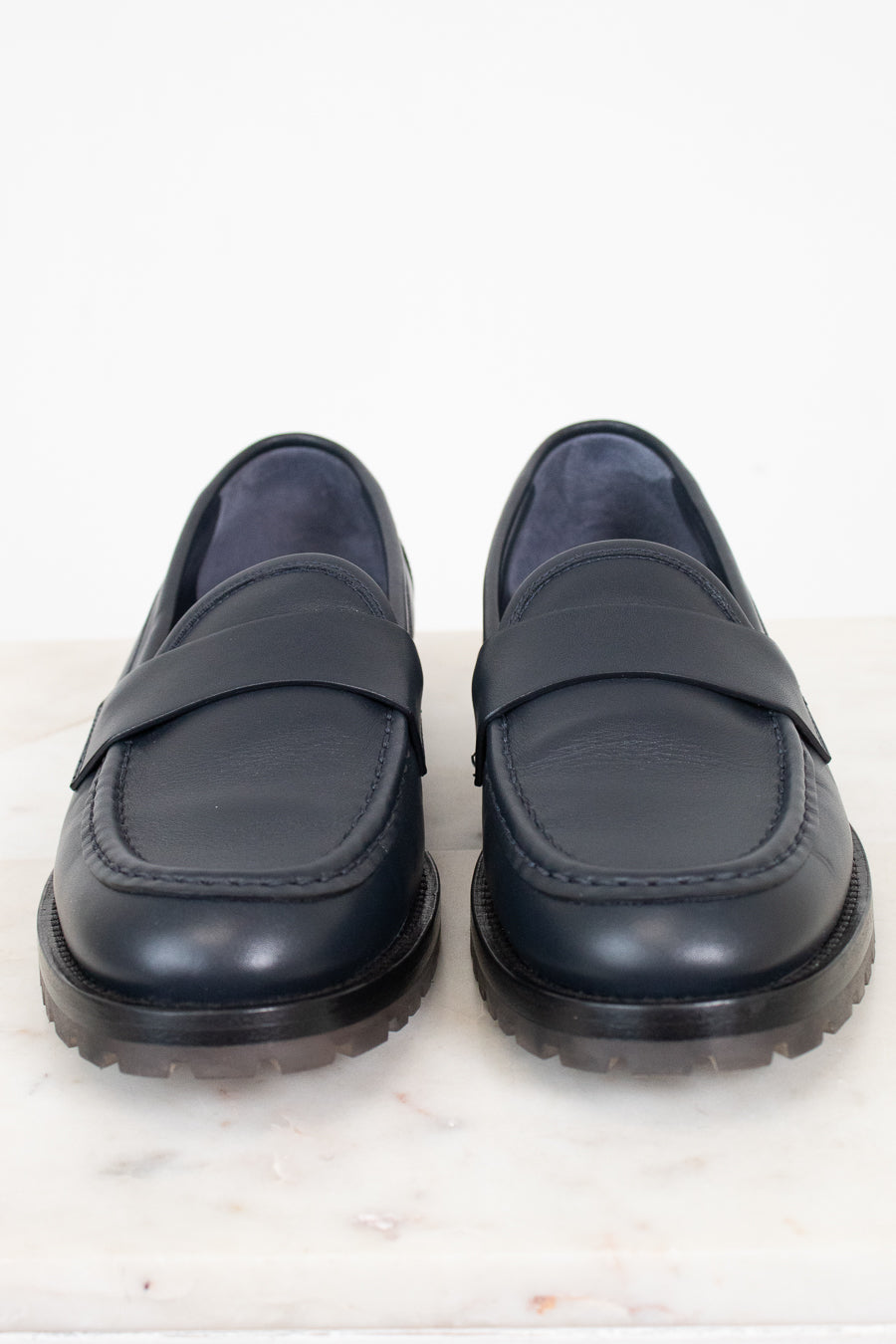 A pair of navy leather penny loafers with thick black soles, displayed side-by-side on a white marble surface against a white background.