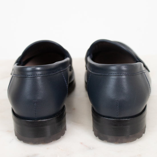 A pair of navy blue leather penny loafers shown from the back, resting on a white marble surface against a white background.