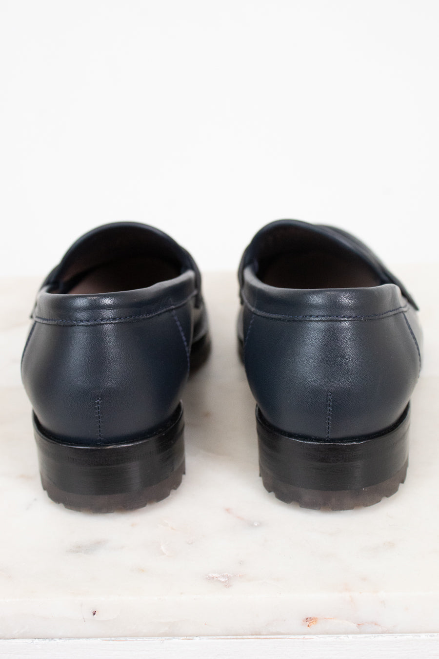 A pair of navy blue leather penny loafers shown from the back, resting on a white marble surface against a white background.