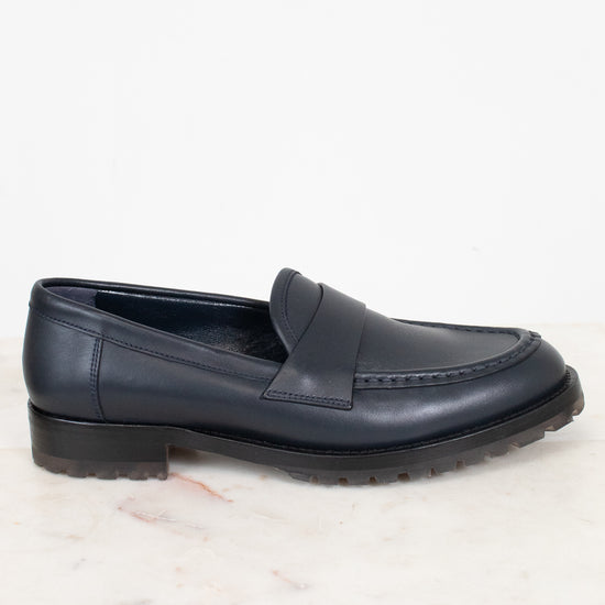 A single dark navy blue leather penny loafer with a low block heel and lug sole, resting on a white marble surface against a white background.