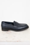 A single dark navy blue leather penny loafer with a low block heel and lug sole, resting on a white marble surface against a white background.