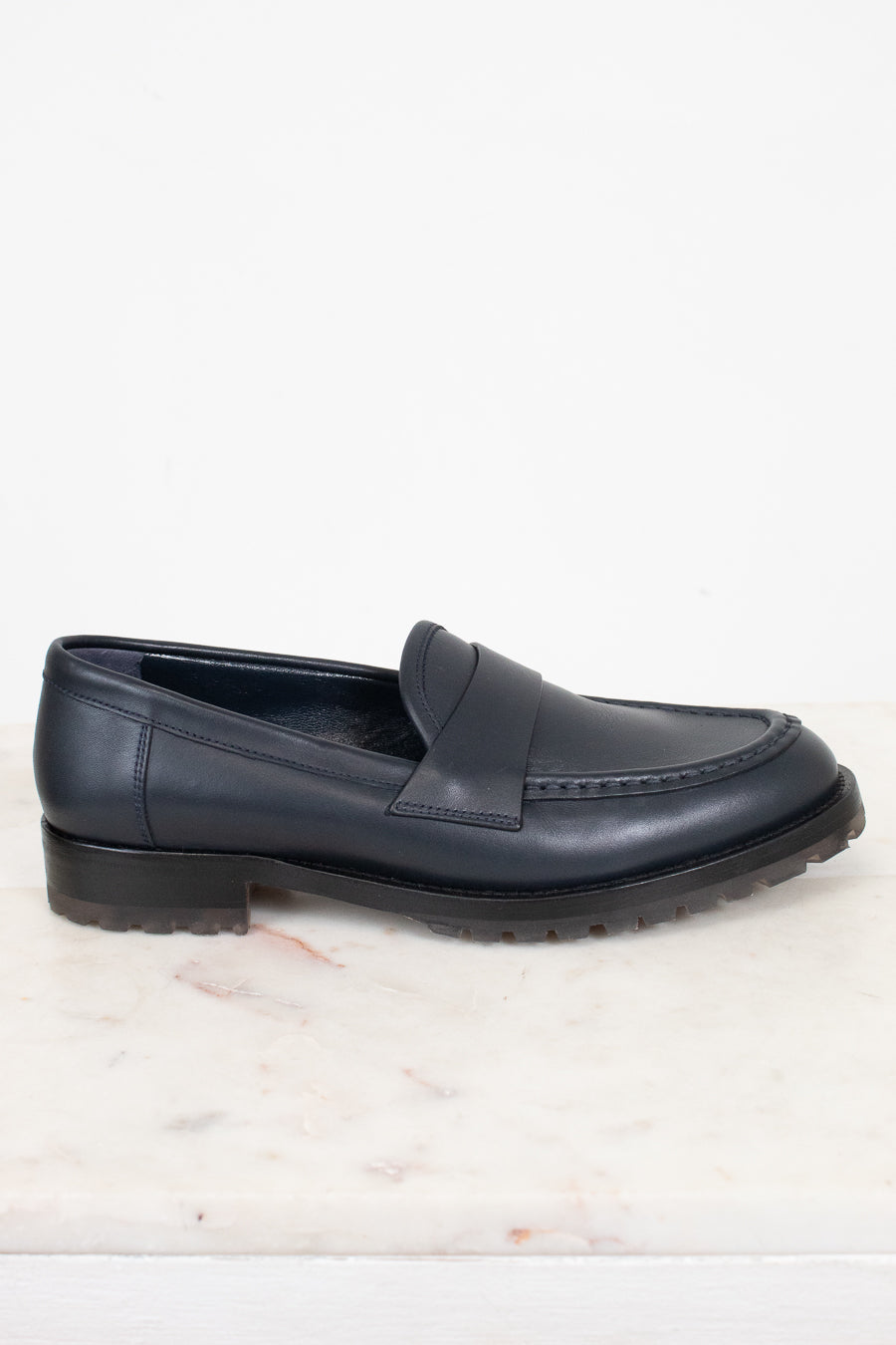 A single dark navy blue leather penny loafer with a low block heel and lug sole, resting on a white marble surface against a white background.