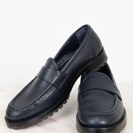 A pair of navy leather penny loafers with chunky black soles, displayed on a light marble surface against a white background.