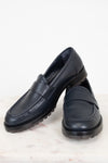A pair of navy leather penny loafers with chunky black soles, displayed on a light marble surface against a white background.