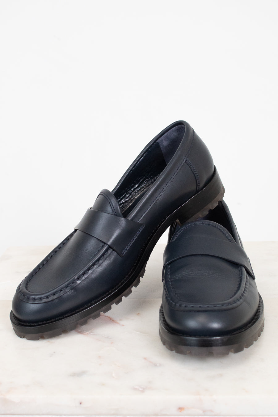 A pair of navy leather penny loafers with chunky black soles, displayed on a light marble surface against a white background.